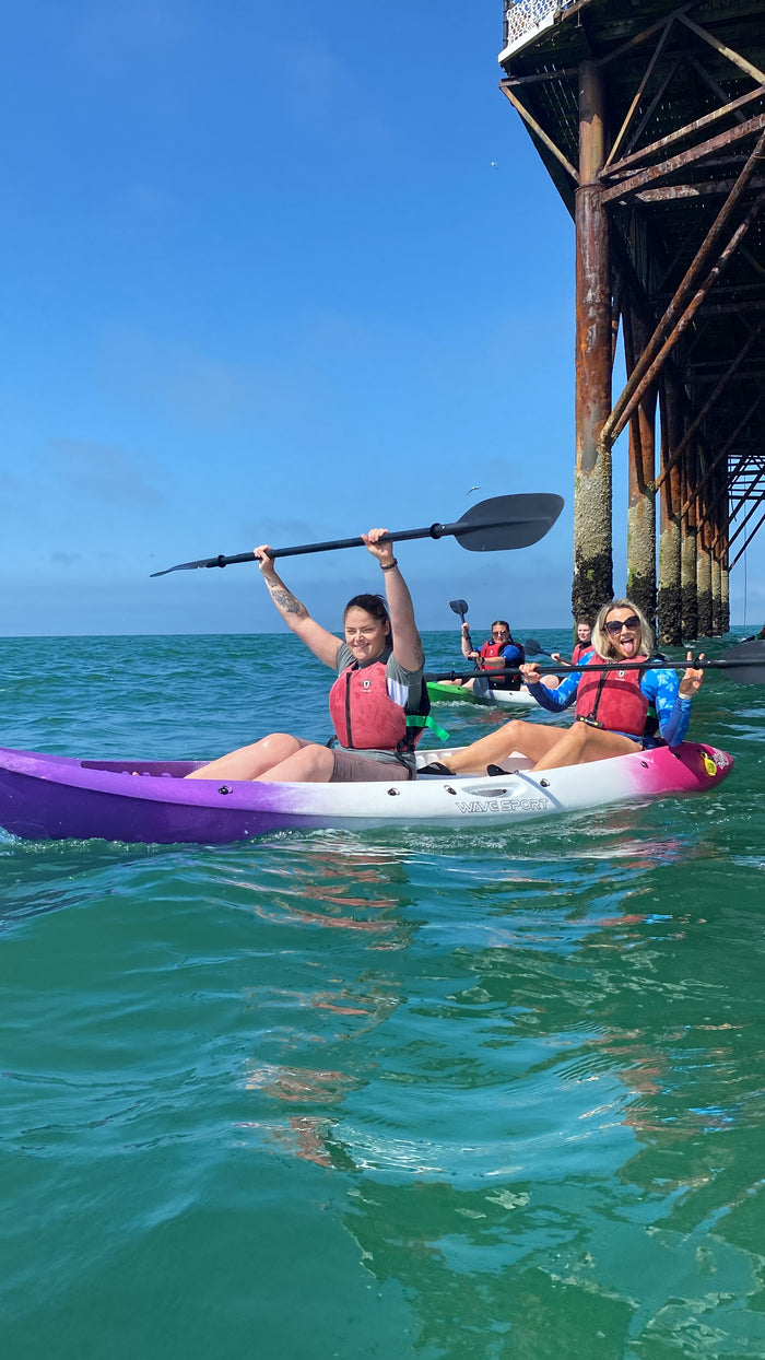Two people kayaking under a pier, with one person paddling and the other sitting in a purple kayak.