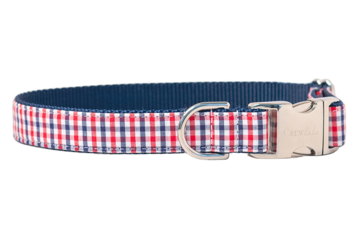 Close-up of a pet collar with a classic red, white, and navy gingham check pattern on the outer layer. The collar is reinforced with navy nylon backing and features a shiny silver buckle clasp with "Crew LaLa" branding, as well as a sturdy metal D-ring.
