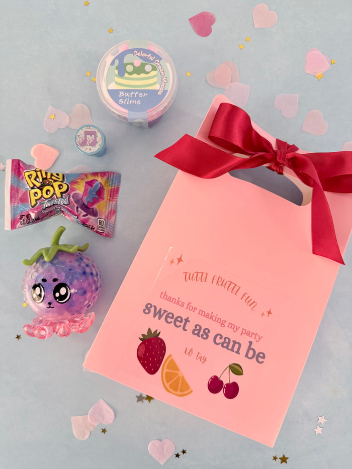 Pink party favor bag with red ribbon, candy, and decorations on a light gray background