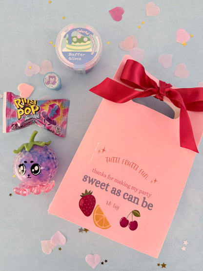 Pink party favor bag with red ribbon, candy, and decorations on a light gray background