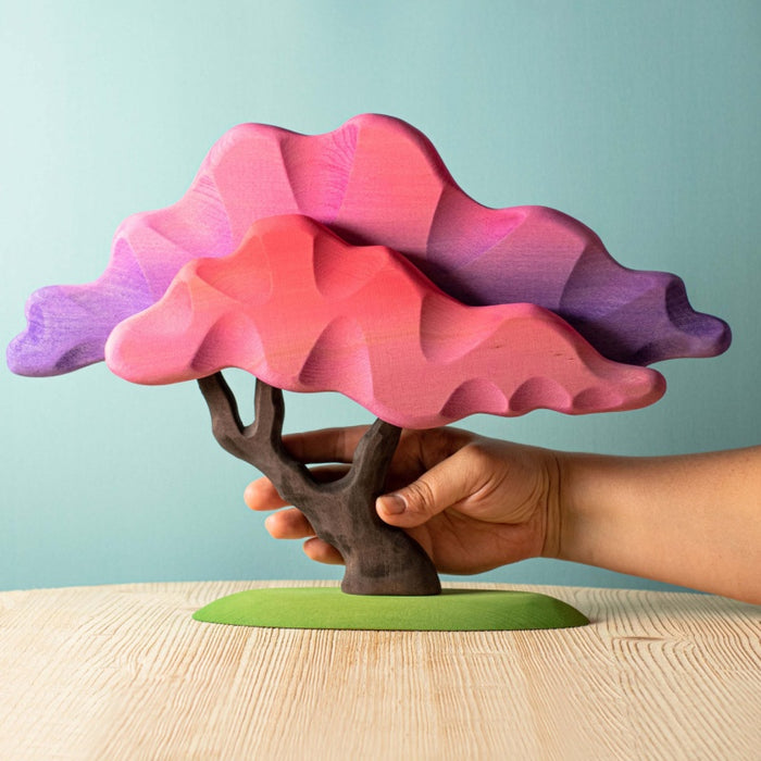 Hand holding a colorful tree-shaped object with a gradient from pink to purple against a light blue background.