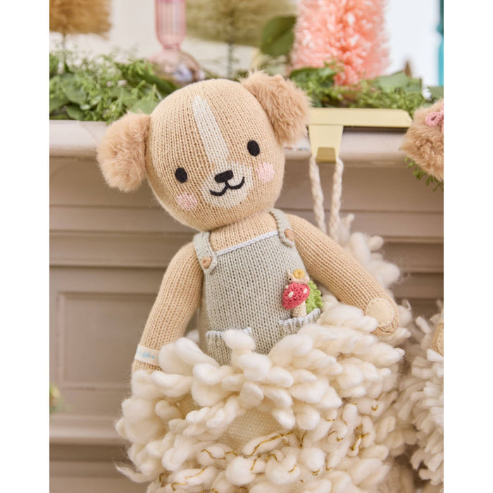 Knitted teddy bear in overalls sitting on a fluffy white surface with a blurred background