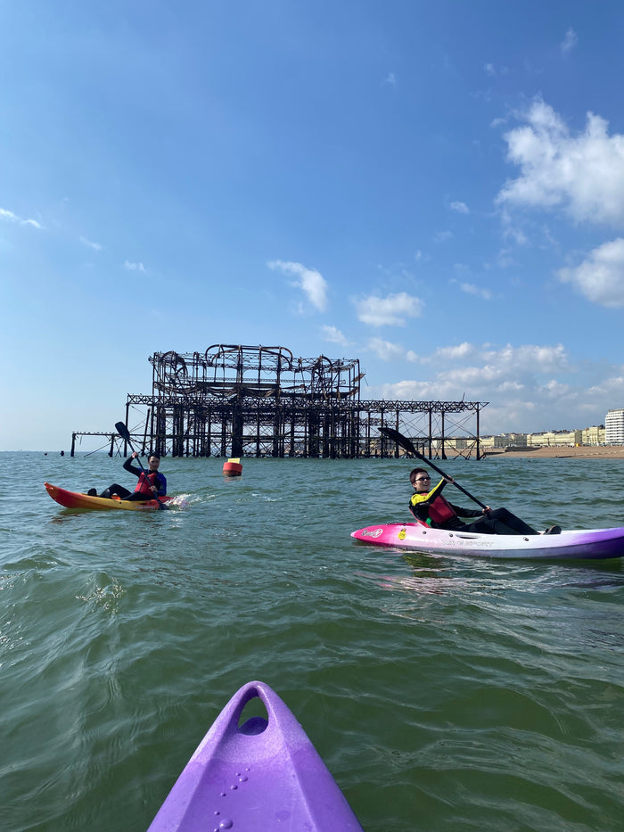 Two people kayaking in open kayaks on the sea, with Brighton Beach and an old pier in the background.