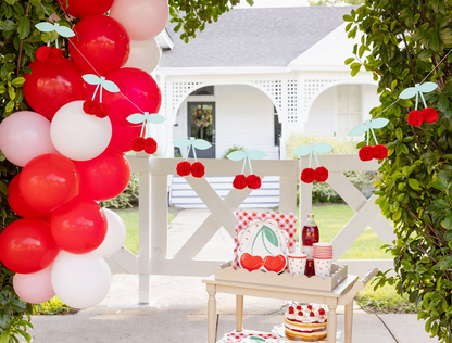 Outdoor setting with red, white, and pink balloons, cherry decorations, and a small table with a cake.