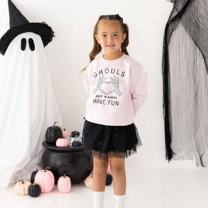 A young girl stands in a Halloween-themed studio setting wearing a light pink sweatshirt with 'Ghouls Just Wanna Have Fun' printed on the front, paired with a black tulle tutu skirt and white socks. She is posed in front of a ghost decoration, pumpkins, and black netting.