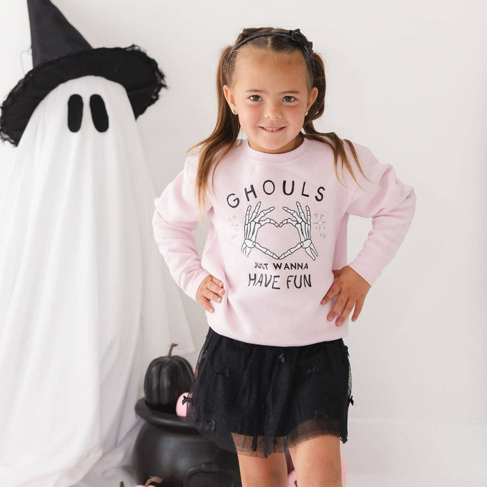 A young girl with brown hair in a black bow stands in a Halloween-themed room wearing a light pink sweatshirt with a black graphic that reads 'GHOULS JUST WANNA HAVE FUN' and a black tulle skirt. To her left is a large white ghost decoration.