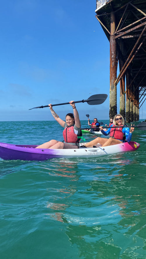 Two people kayaking under a pier, with one person paddling and the other sitting in a purple kayak.
