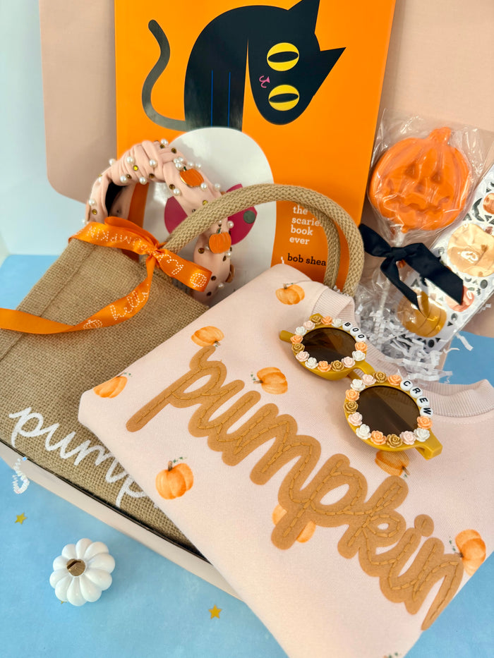 A styled gift box arrangement featuring a beige tote bag with an orange ribbon, a cream sweatshirt printed with 'pumpkin' in tan letters and small pumpkin motifs, novelty sunglasses with jeweled frames, a small pumpkin ornament, and an orange book or card with a black cat illustration.