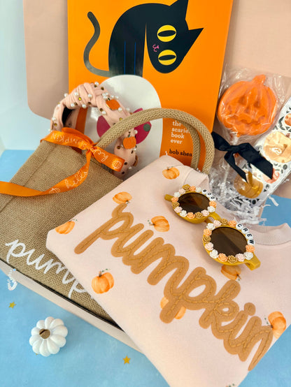 A styled gift box arrangement featuring a beige tote bag with an orange ribbon, a cream sweatshirt printed with 'pumpkin' in tan letters and small pumpkin motifs, novelty sunglasses with jeweled frames, a small pumpkin ornament, and an orange book or card with a black cat illustration.