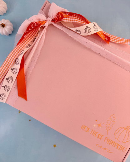 A pink gift box with a personalized name printed in gold on the front, tied with a decorative ribbon featuring orange gingham and white ribbon with pumpkin illustrations. The box is photographed on a light blue background with small decorative pumpkins in the corner.