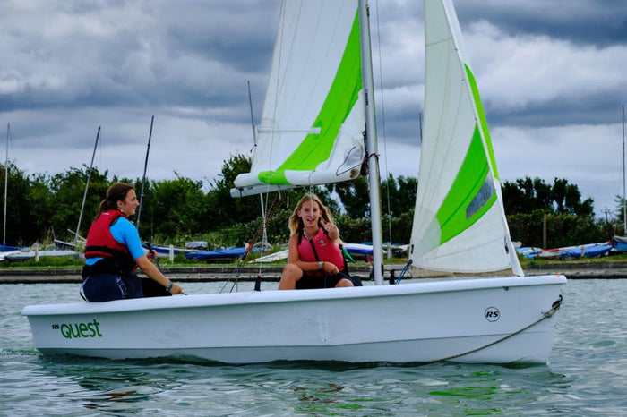 Two people sailing a dinghy with bright green and white sails on a calm lake under cloudy skies.