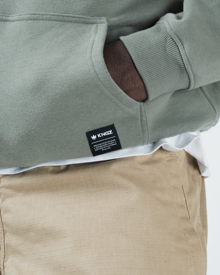Dark forest green BJJ hoodie bottom hem closeup of woven Kingz crown label tag and kangaroo pocket opening