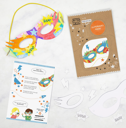 A colorful superhero mask with yellow straps and a cardboard base, alongside a kraft envelope with an instruction card and glue dots.