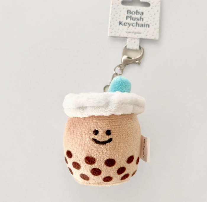 A small plush keychain in the shape of a boba milk tea cup with a white and brown color scheme, featuring a smiling face, a white pom-pom on top, and a blue-patterned surface.