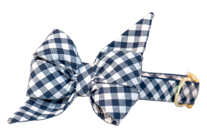 Close-up of a plaid dog collar with an attached matching feminine belle bow featuring two long, pointed tails. The belle bow is made from soft but structured cotton fabric in a classic navy and white check pattern. The collar includes detailed topstitching for added durability and is finished with an adjustable gold metal buckle and matching D-ring, ideal for leash attachment or charm accessories.