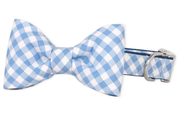Close-up of a coordinated plaid dog collar and bow tie in periwinkle and white. The crisp, fabric-covered collar features a centered bow tie with neatly pleated sides, a sturdy D-ring, and a silver buckle clasp, offering a charming yet practical design for everyday wear or special occasions.