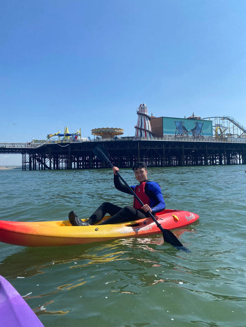 A person kayaking in a red and yellow kayak on calm waters with Brighton Beach and a pier in the background.