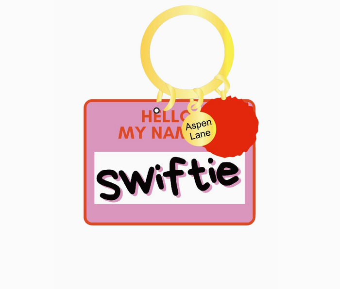 An acrylic keychain and backpack charm with 'Hello my name is Taylor Swift' printed on it, featuring a pink background, yellow ring, and a red pom-pom.