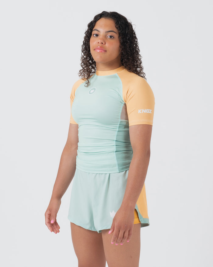 Kingz Terra Women's Rashguard Green Edition - Full Body 3/4 Front Left Side View Female Athlete Short Sleeve Mint Light Green Body Yellow Peach Raglan Sleeves Crown Logo KINGZ White Sleeve Text Matching Mint Hybrid Shorts Women's BJJ [color:Light Green]