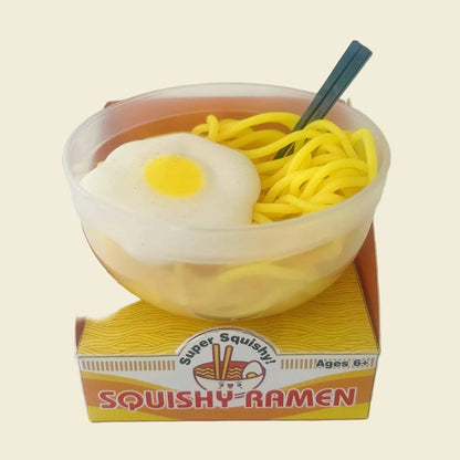 A miniature toy ramen bowl labeled 'Squishy Ramen' sitting on its packaging box. The toy features yellow noodles, a white fried egg with a yellow yolk, and a dark green chopstick in a clear plastic bowl.